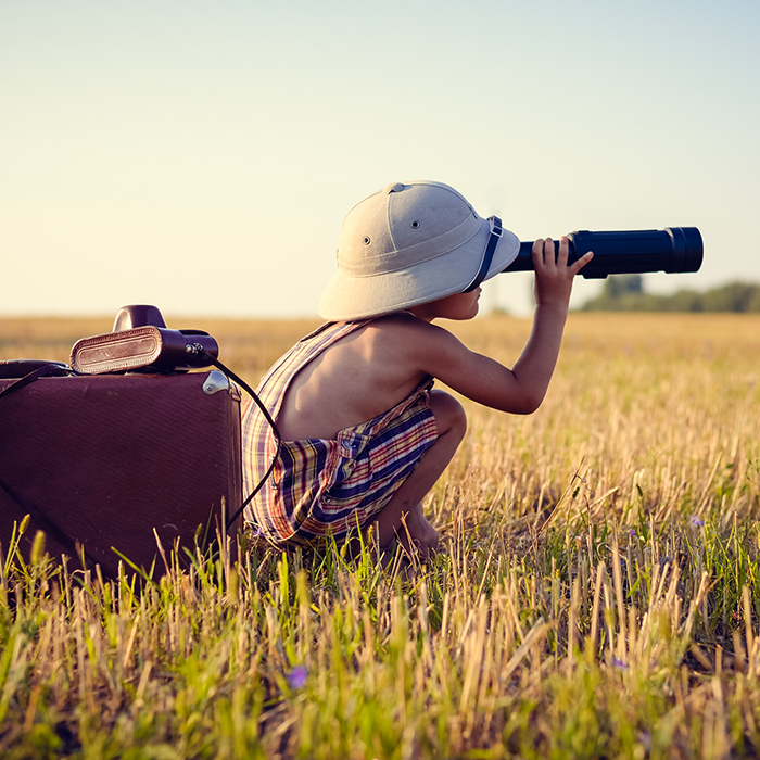 Young boy squatting in field looking in the distance through telescope